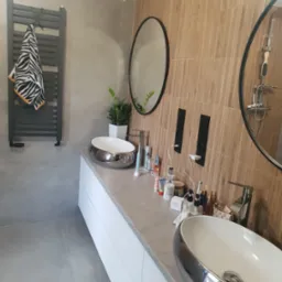 Nowoczesna łazienka z dwoma owalnymi umywalkami w srebrnej obudowie, białymi szafkami pod umywalkami, drewnopodobnymi płytkami na ścianie z dwoma okrągłymi lustrami i czarnymi gniazdkami...