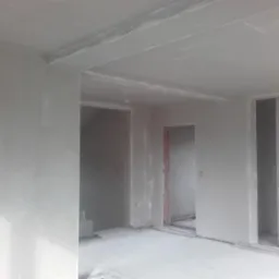 Surowe wnętrze w trakcie prac wykończeniowych, ściany pokryte świeżym tynkiem, widoczne otwory drzwiowe z częściowo wykończonymi ościeżnicami, przewody elektryczne na podłodze.