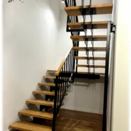 Nowoczesne schody z drewnianymi stopniami i czarną, metalową konstrukcją wsporczą, zamontowane przy białej ścianie, z widoczną drewnianą podłogą i fragmentem balustrady z drewnianą poręczą.