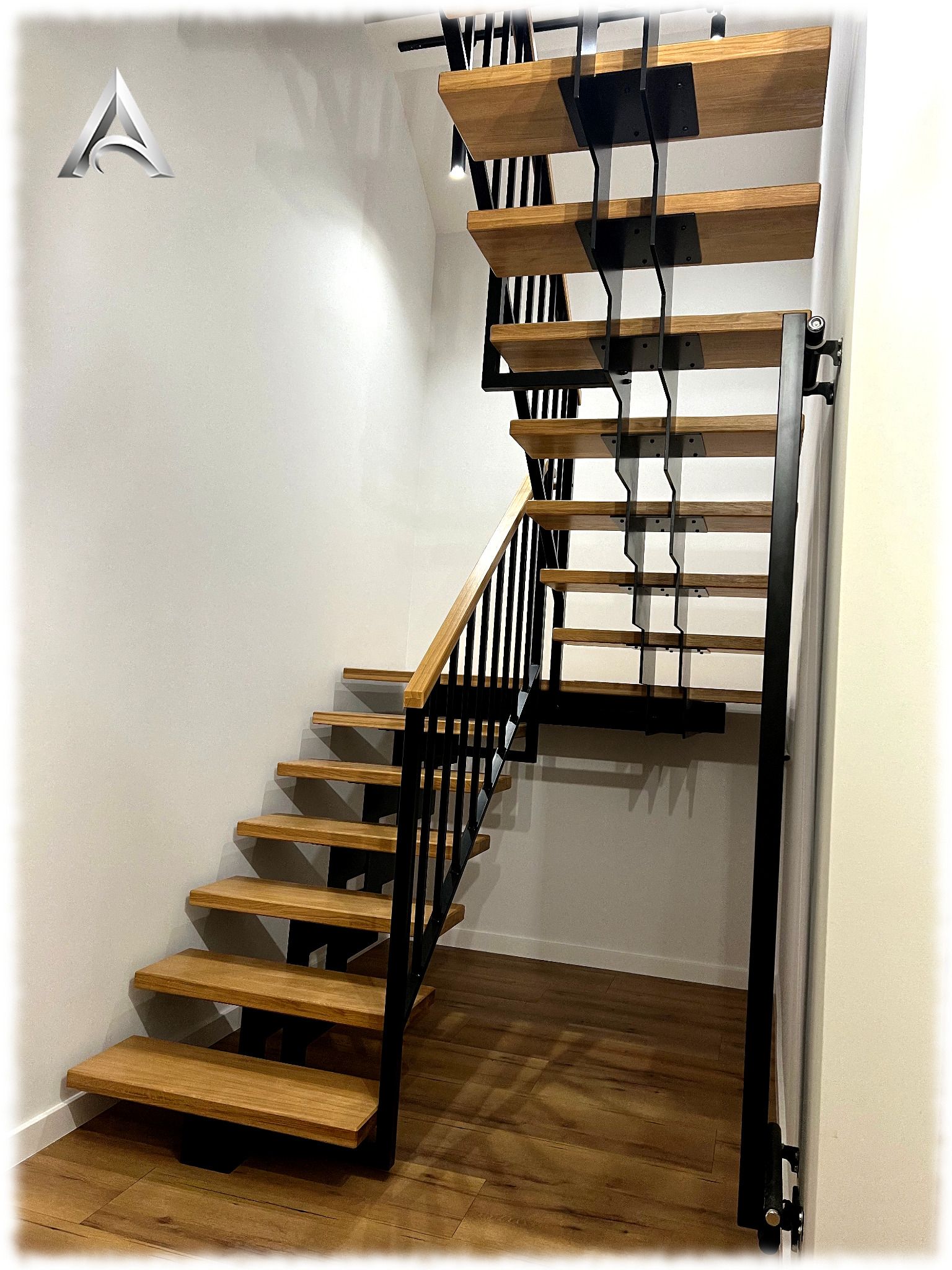 Nowoczesne schody z drewnianymi stopniami i czarną, metalową konstrukcją wsporczą, zamontowane przy białej ścianie, z widoczną drewnianą podłogą i fragmentem balustrady z drewnianą poręczą.
