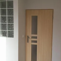 Zamknięte drzwi z szybą w drewnianej okleinie, zamontowane w jasnym wnętrzu z podłogą imitującą drewno i szklaną ścianką działową z luksferów.