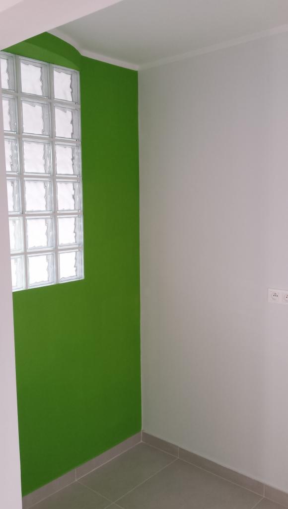 Wnętrze po remoncie: ściana w odcieniach zieleni z oknem z luksferów, biała ściana z gniazdkami elektrycznymi i szara podłoga z płytek. Nowoczesne wykończenie.