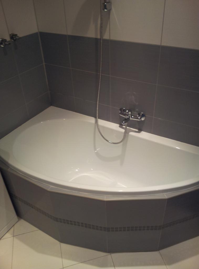 Biała, narożna wanna z szarą obudową z płytek i chromowaną armaturą. Ściany wyłożone szarymi płytkami do połowy wysokości.