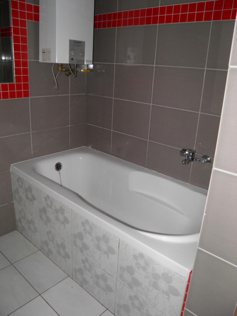 Wykończona łazienka z szarą glazurą, białą wanną obudowaną płytkami w kwiaty i piecykiem gazowym. Czerwony akcent w postaci dekoracyjnej listwy z płytek.