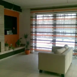 Jasny salon z beżową sofą, kominkiem i oknami przesłoniętymi roletami rzymskimi w poziome pasy pomarańczowe, białe i szare.