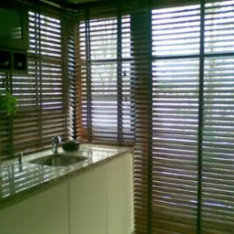 Kuchnia z kamiennym blatem i drewnianymi żaluzjami poziomymi w oknach, widok na zewnątrz przez szpary między lamelami.