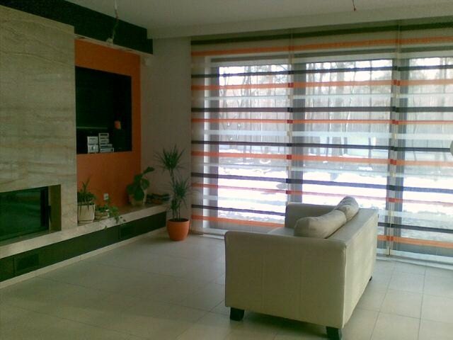 Jasny salon z beżową sofą, kominkiem i oknami przesłoniętymi roletami rzymskimi w poziome pasy pomarańczowe, białe i szare.