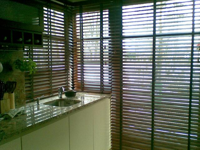 Kuchnia z kamiennym blatem i drewnianymi żaluzjami poziomymi w oknach, widok na zewnątrz przez szpary między lamelami.