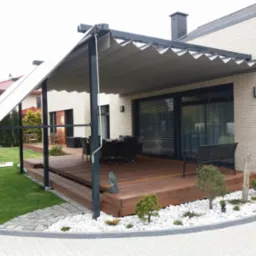Nowoczesna pergola z rozsuwanym zadaszeniem materiałowym w kolorze szarym, metalowa konstrukcja, drewniany taras, białe kamienie dekoracyjne, zadbany trawnik i elementy małej architektury ogrodowej.
