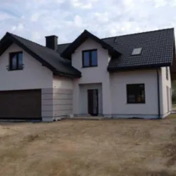 Nowoczesny, dwukondygnacyjny dom z garażem i czarnym dachem, z elewacją w odcieniach bieli i szarości, na działce z widoczną świeżo nawiezioną ziemią.