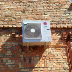 Klimatyzator LG Inverter V zamontowany na ceglanej ścianie budynku, widoczne rury instalacyjne i cień padający na mur.