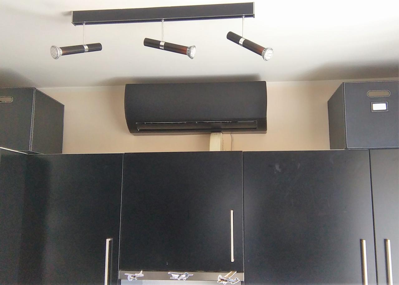 Czarny klimatyzator ścienny zamontowany nad ciemnymi szafkami kuchennymi, oświetlony trzema reflektorami na listwie.