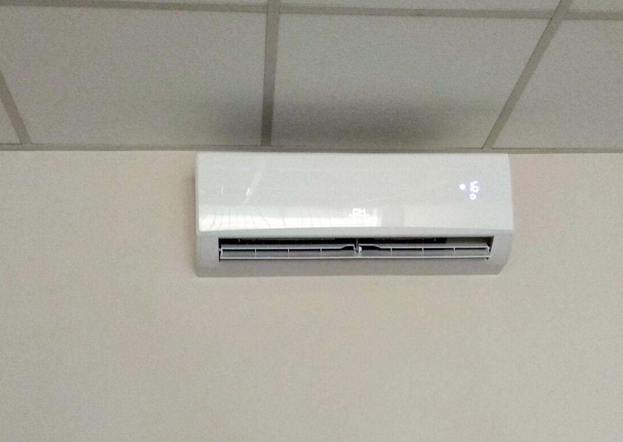 Biała jednostka klimatyzacji, zamontowana na ścianie w pomieszczeniu w Mosinie, widoczny wyświetlacz z temperaturą i logo producenta, tło stanowi sufit kasetonowy.