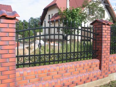 Czarne, kute ogrodzenie z dekoracyjnymi elementami, osadzone na niskim murku z czerwonej cegły i słupkach z ceglaną nakładką, na tle domu jednorodzinnego.