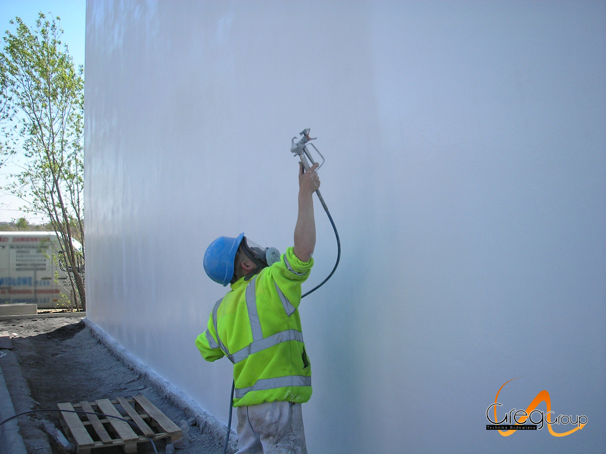 Pracownik w kasku i masce maluje natryskowo dużą, białą ścianę zewnętrzną, widoczny pistolet natryskowy i fragment palety na ziemi.