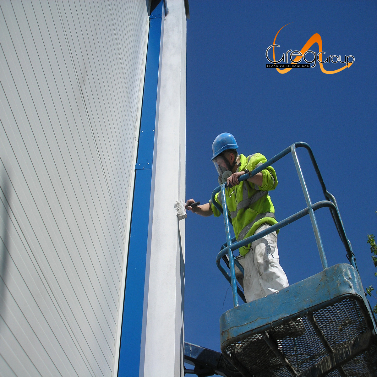 Pracownik w kasku i masce maluje białą farbą filar budynku z podnośnika koszowego, widoczna elewacja z szarych paneli i niebieski element konstrukcyjny.