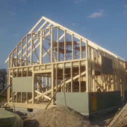 Konstrukcja drewniana domu w trakcie budowy, widoczne elementy szkieletu, częściowo obłożone płytami, z rusztowaniami wewnątrz, na tle błękitnego nieba i fragmentu innego budynku.