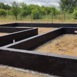 Zabezpieczone hydroizolacją fundamenty domu jednorodzinnego, widoczne wystające pręty zbrojeniowe i otwory wentylacyjne, teren przygotowany pod dalsze prace.