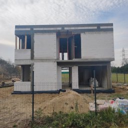 KAST-BUD Usługi budowlane Tomasz Kot - Dwukondygnacyjny dom w stanie surowym otwartym, zbudowany z białych bloczków, widoczny przez ogrodzenie z siatki. Na pierwszym planie piasek i roślinność.