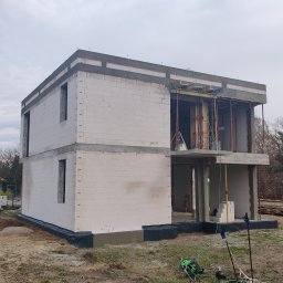 KAST-BUD Usługi budowlane Tomasz Kot - Dwukondygnacyjny dom w trakcie budowy, ściany z białych bloczków, widoczne zbrojenia i szalunki, teren budowy z narzędziami. Ujęcie z dołu, pochmurne niebo.