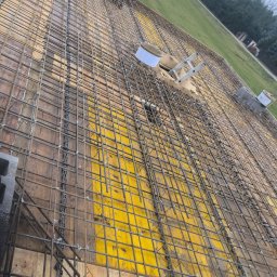 KAST-BUD Usługi budowlane Tomasz Kot - Siatka zbrojeniowa na deskowaniu, widok z góry. Żółte płyty szalunkowe, stalowe pręty połączone drutem. W tle trawa i budynek. Zbrojenie stropu.