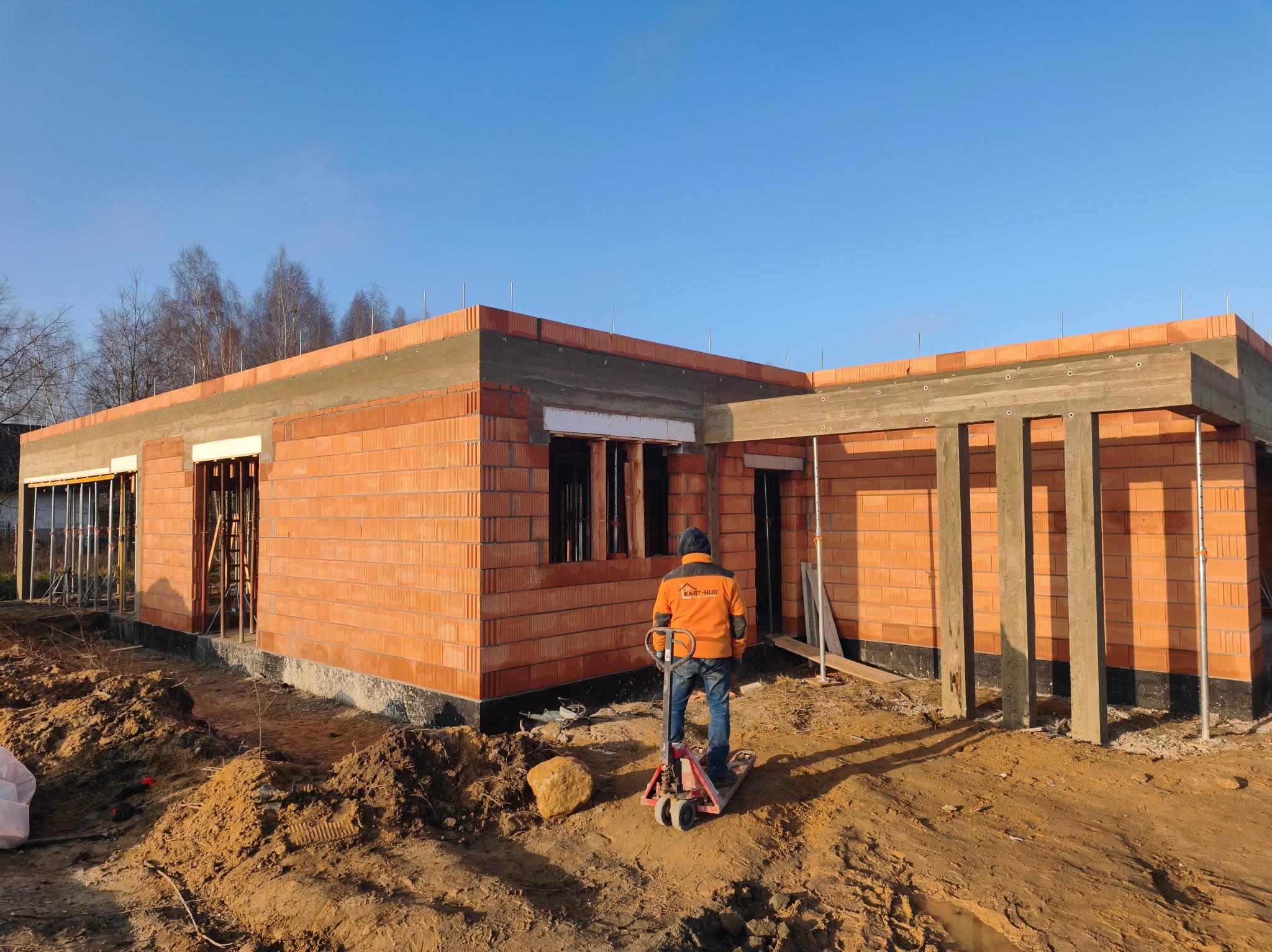 Budowa domu z czerwonej cegły: murarz z wózkiem transportowym na tle wznoszonej konstrukcji, widoczne betonowe nadproża i elementy zbrojenia. Słoneczny dzień.