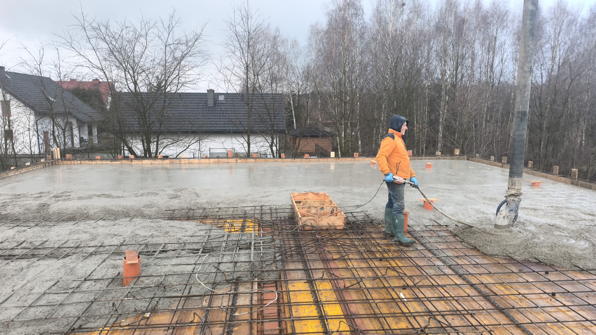 Wylewanie betonu na zbrojenie fundamentów domu jednorodzinnego. Pracownik w pomarańczowej kurtce rozprowadza beton. W tle dom i drzewa.
