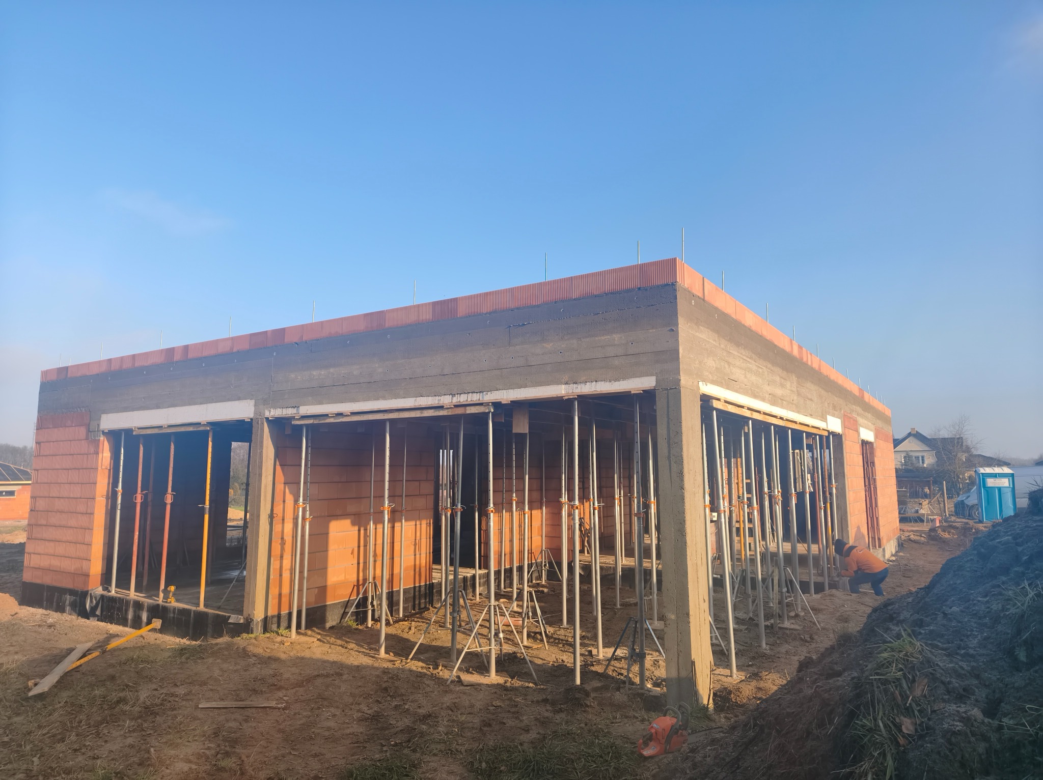 Budowa domu jednorodzinnego w stanie surowym otwartym: ściany z czerwonej cegły, betonowe słupy, stalowe podpory, widoczny pracownik w pomarańczowej kurtce.