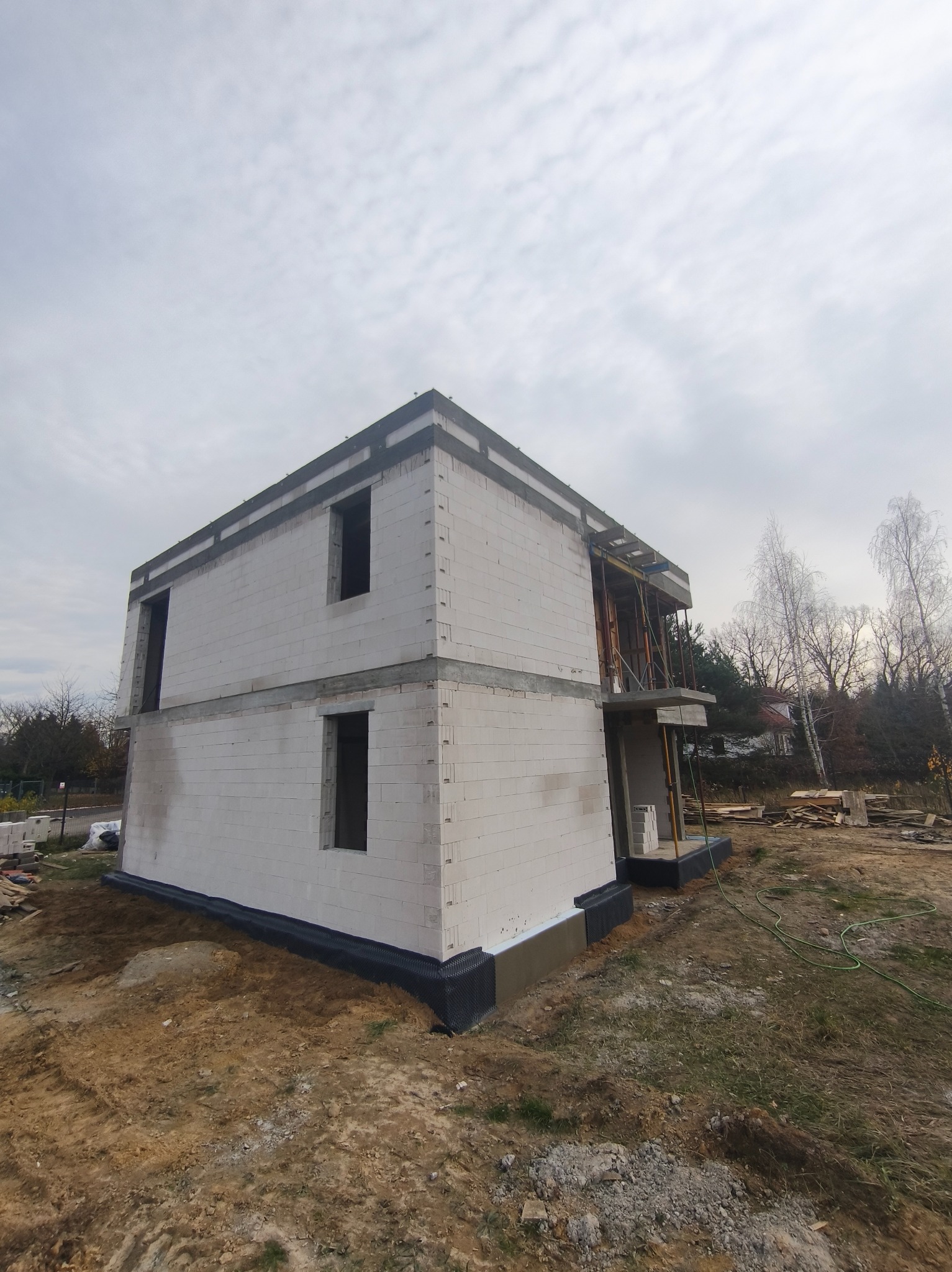 Surowy, dwukondygnacyjny dom z białego bloczka, w trakcie budowy. Widoczne szalunki na balkonie, teren wokół w stanie surowym. Pochmurne niebo w tle.