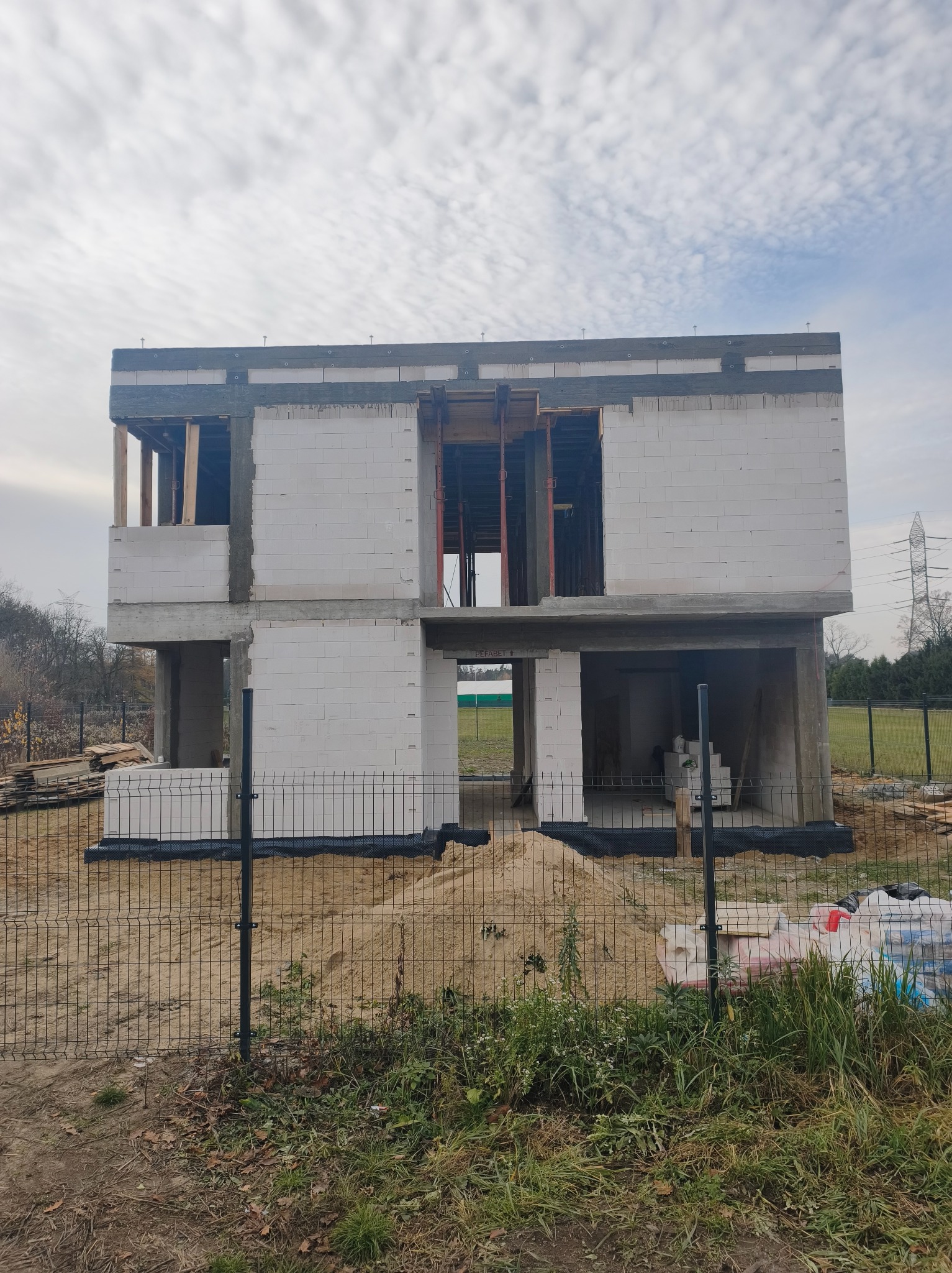 Dwukondygnacyjny dom w stanie surowym otwartym, zbudowany z białych bloczków, widoczny przez ogrodzenie z siatki. Na pierwszym planie piasek i roślinność.