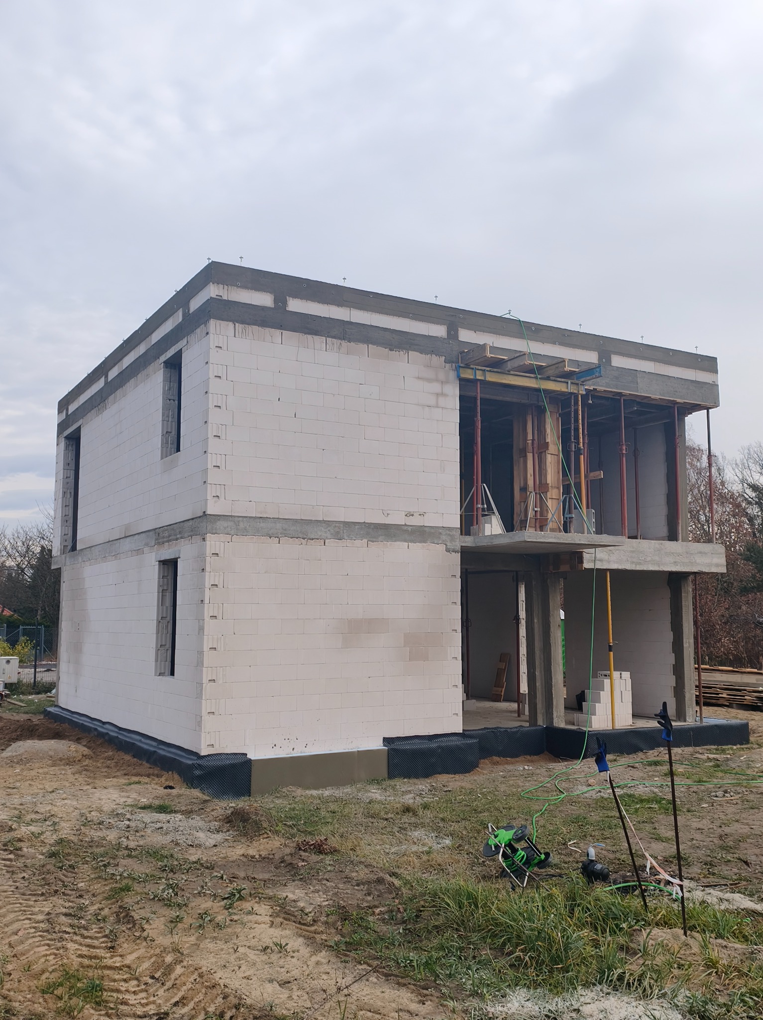 Dwukondygnacyjny dom w trakcie budowy, ściany z białych bloczków, widoczne zbrojenia i szalunki, teren budowy z narzędziami. Ujęcie z dołu, pochmurne niebo.