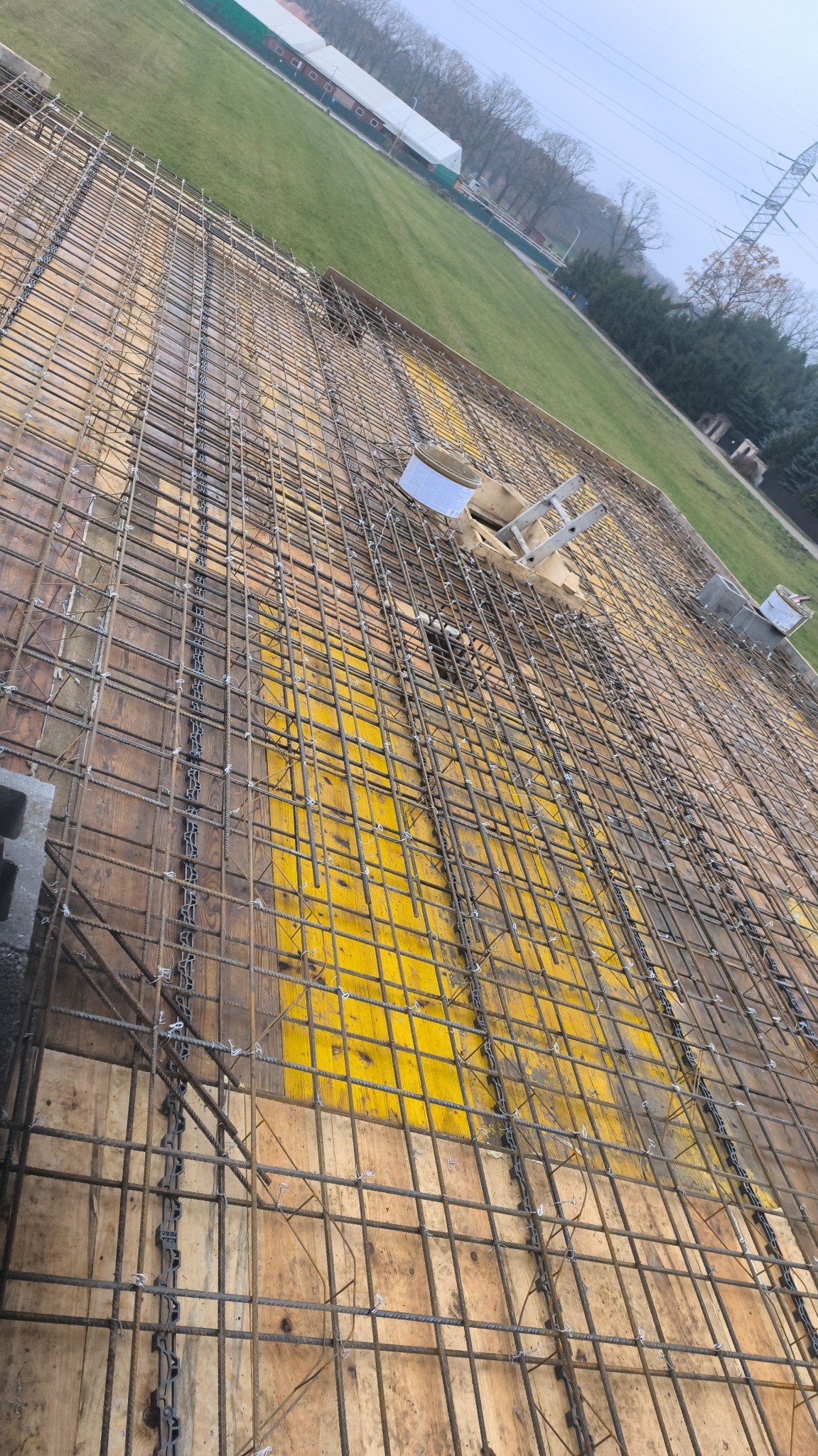 Siatka zbrojeniowa na deskowaniu, widok z góry. Żółte płyty szalunkowe, stalowe pręty połączone drutem. W tle trawa i budynek. Zbrojenie stropu.