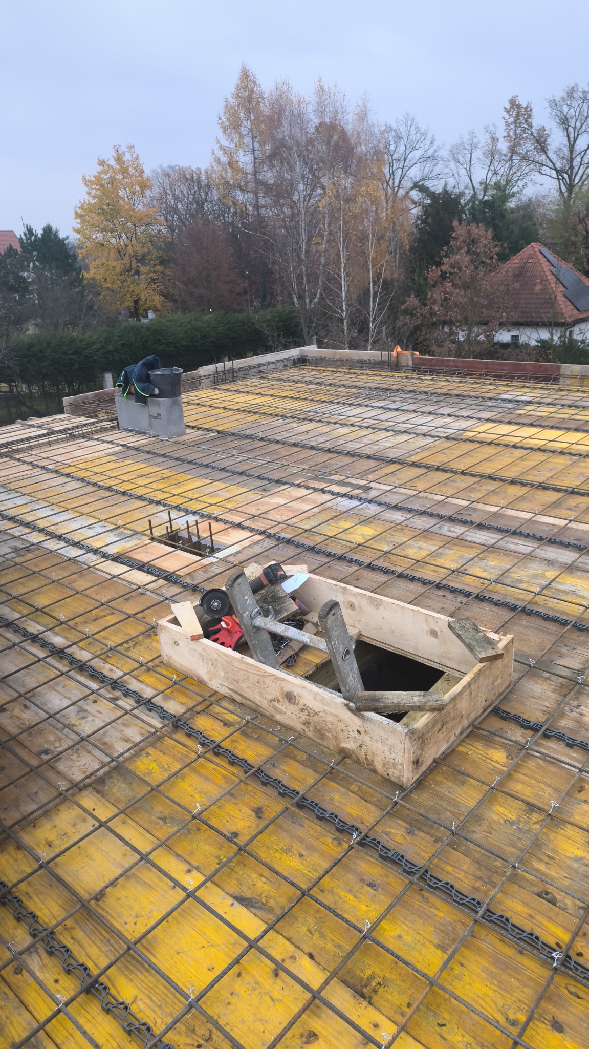 Żółty szalunek stropowy z siatką zbrojeniową, drewniana skrzynia z narzędziami i drabina w centrum kadru. W tle drzewa i fragment dachu.
