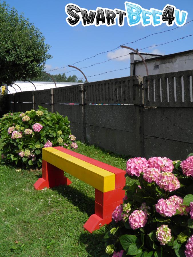 Ławka ogrodowa SmartBee4u w Koszalinie, pomalowana na czerwono i żółto, ustawiona na trawniku pomiędzy kwitnącymi hortensjami na tle betonowego ogrodzenia z drutem kolczastym.