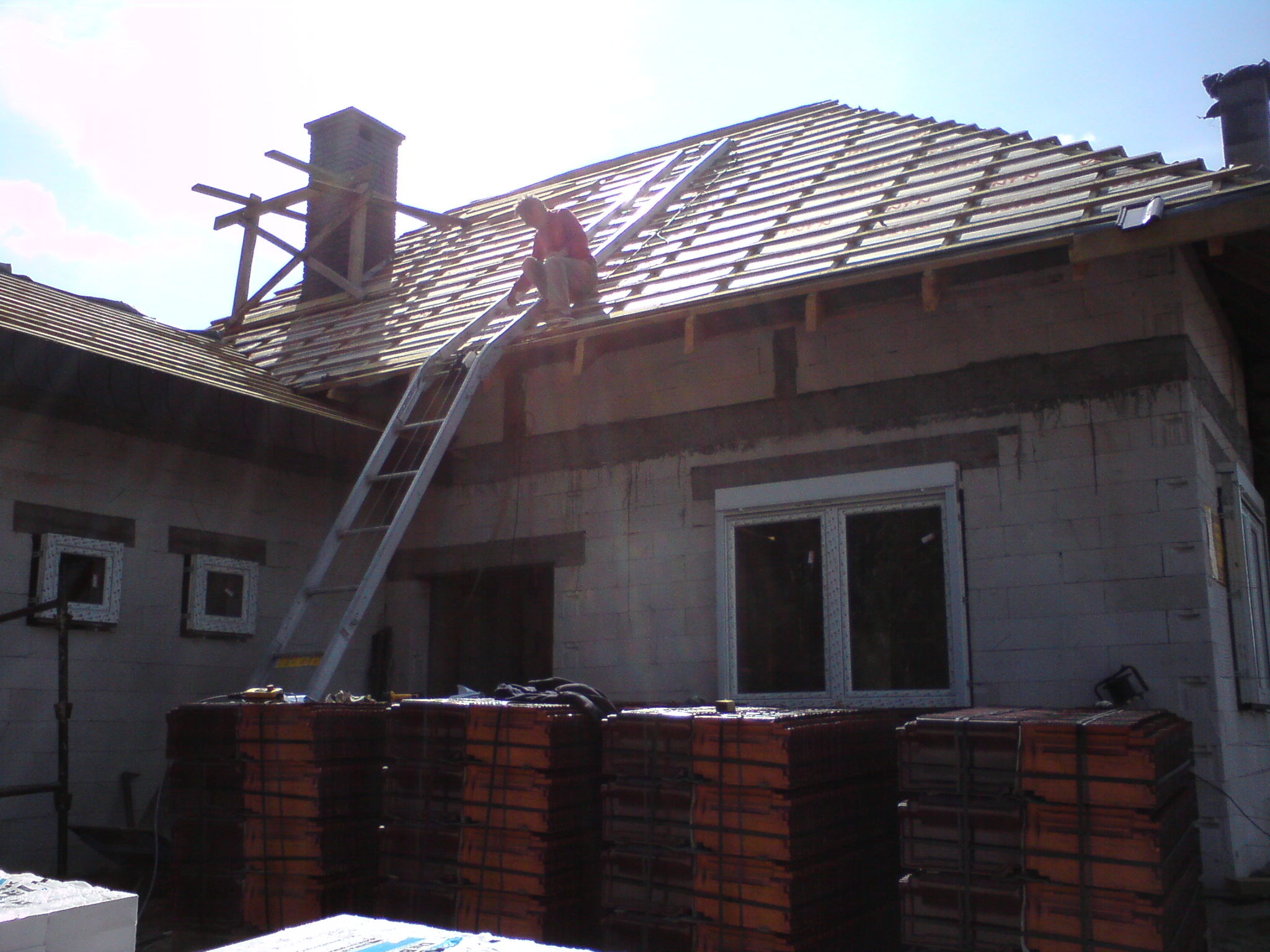 Ceglasta dachówka ułożona na konstrukcji dachu domu w trakcie budowy; pracownik na drabinie montuje elementy konstrukcyjne w pobliżu komina; na pierwszym planie palety z dachówkami i pustaki.