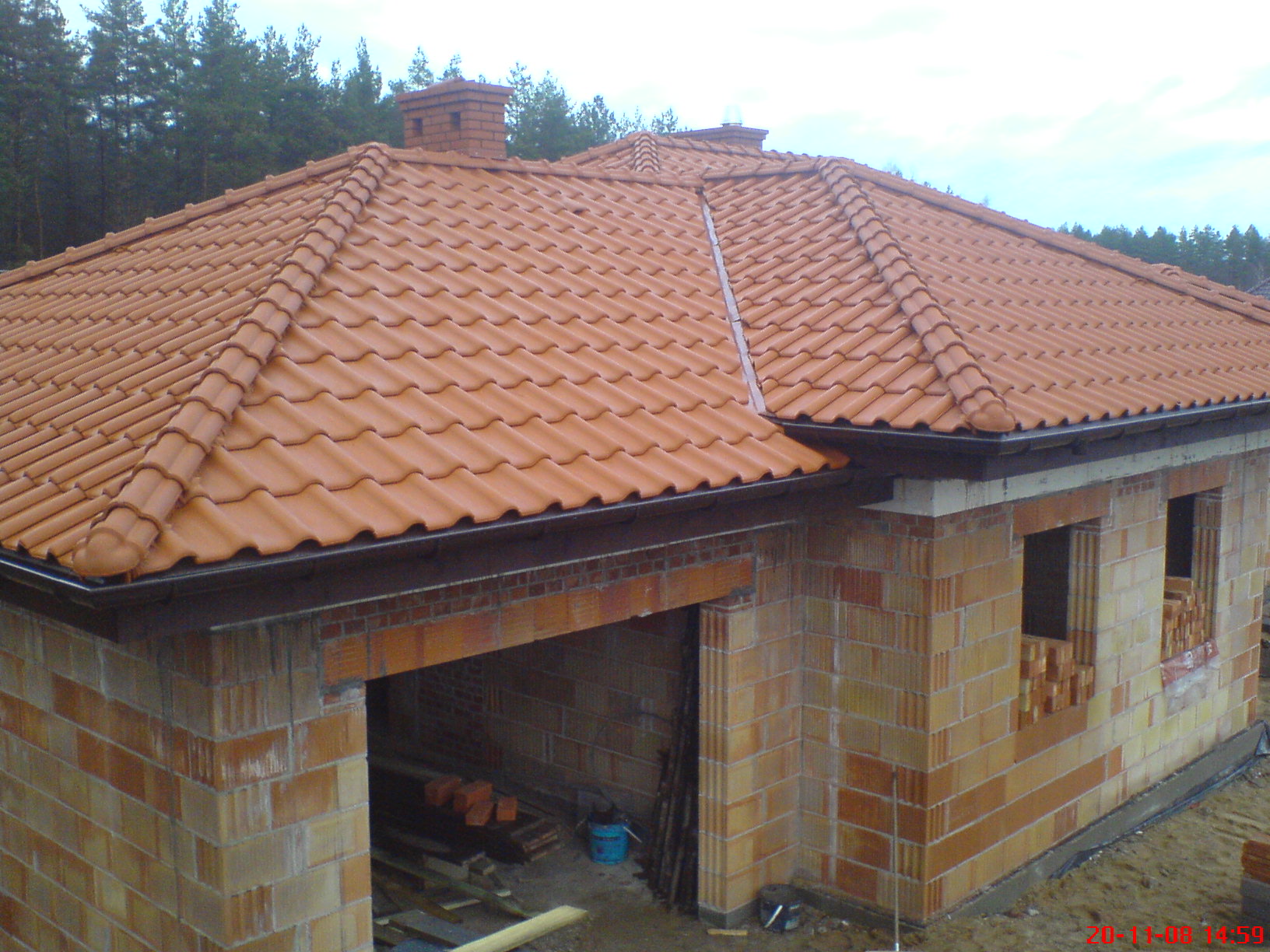 Niedokończony budynek z pomarańczową dachówką, widoczne niezamurowane okna i garaż z materiałami budowlanymi w środku, las w tle.