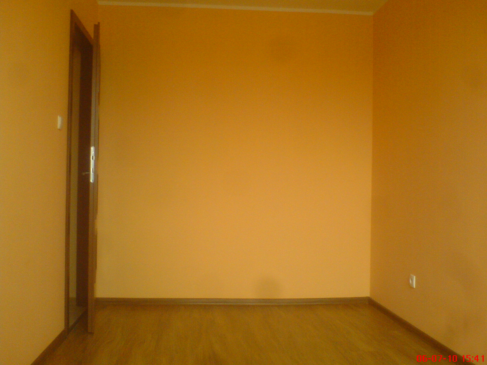 Pomarańczowe ściany i drewnopodobna podłoga w pustym pokoju po remoncie, widoczny otwarty fragment drzwi.