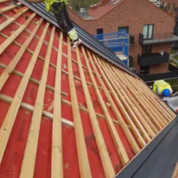 Pracownik w kasku i kamizelce odblaskowej montuje metalowe elementy na drewnianej konstrukcji dachu pokrytej czerwoną membraną, w tle budynki mieszkalne.