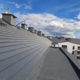 Nowoczesny dach z szarej dachówki ceramicznej na budynku wielorodzinnym, widoczne kominy, błękitne niebo z chmurami w tle.