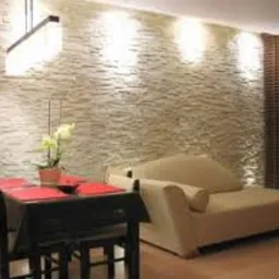 Ściana z dekoracyjną glazurą w odcieniach beżu i brązu, oświetlona punktowymi lampami, tworząca przytulne wnętrze w mieszkaniu w Warszawie. Na pierwszym planie stół z krzesłami i sofa.