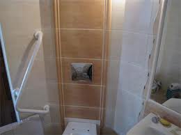 Beżowo-kremowa glazura na ścianie w łazience z widocznym przyciskiem spłuczki, uchwytem dla niepełnosprawnych i fragmentem lustra. Widoczne niedoskonałości montażu.