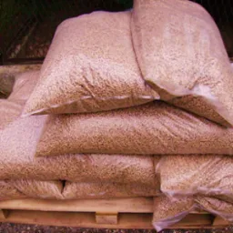 Stos worków z granulatem na drewnianej palecie, widoczne przez przezroczyste opakowanie, z zewnątrz na podłożu żwirowym i fragmentem trawnika.