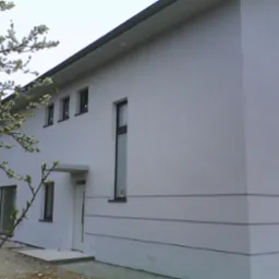 Nowoczesny dom jednorodzinny z jasną elewacją, widoczne trzy cienkie poziome linie dekoracyjne, świeżo otynkowana ściana, drzewo z kwitnącymi gałęziami na pierwszym planie, teren wokół domu jeszcze...