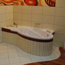 Wanna obudowana panelami w łazience z beżowymi płytkami i abstrakcyjnym wzorem na górze ścian.