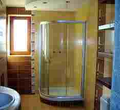 Kabina prysznicowa z zaokrąglonymi drzwiami w narożniku łazienki. Widoczna brązowa glazura na ścianie z oknem oraz żółta ściana obok kabiny. Ujęcie z poziomu podłogi.