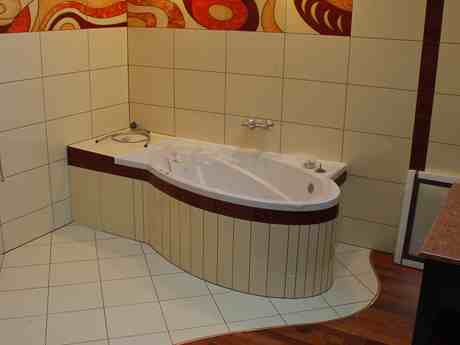 Wanna obudowana panelami w łazience z beżowymi płytkami i abstrakcyjnym wzorem na górze ścian.