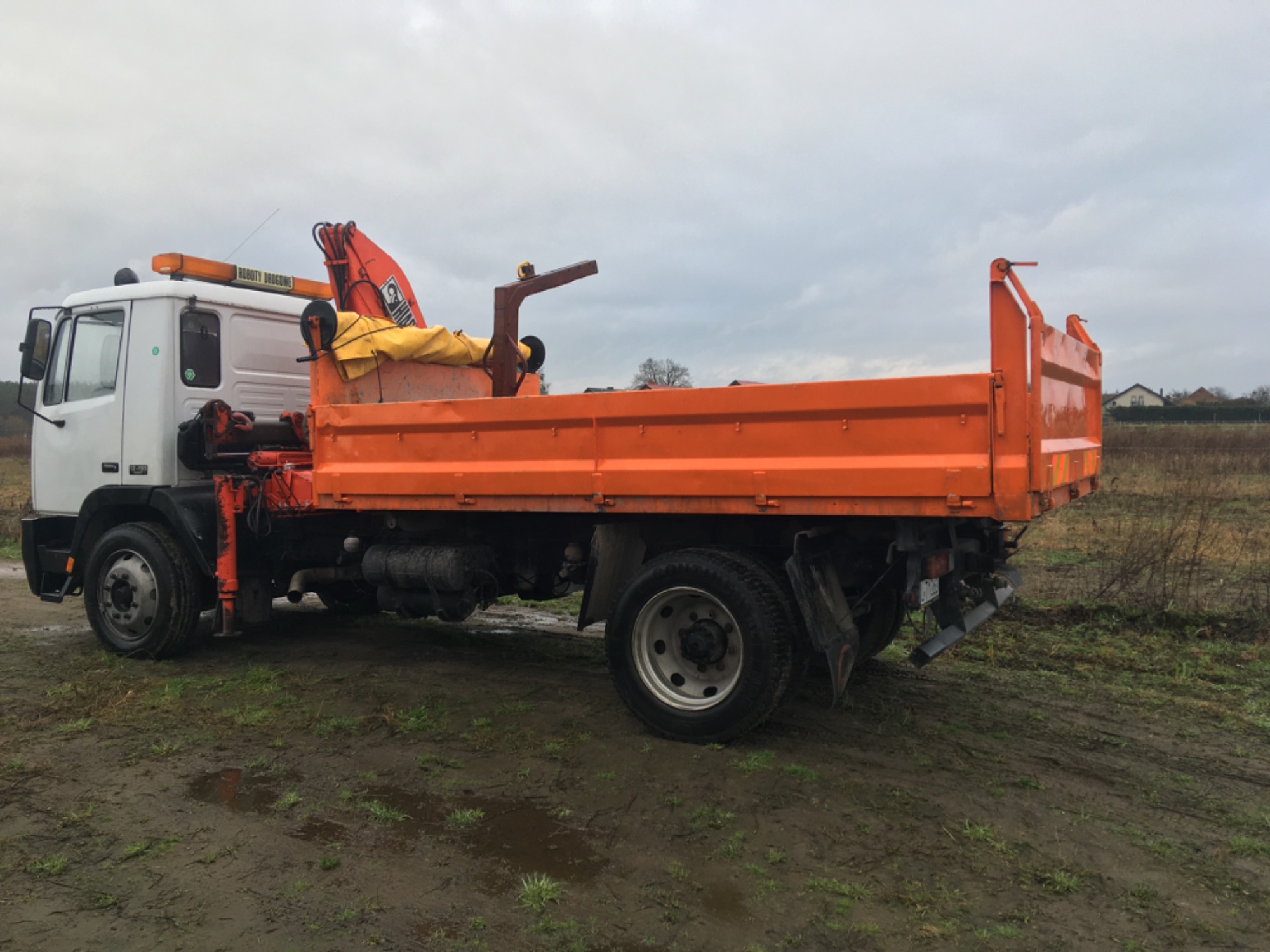 Pomarańczowa ciężarówka z dźwigiem Hiab na błotnistym terenie, widok z boku, pochmurne niebo.
