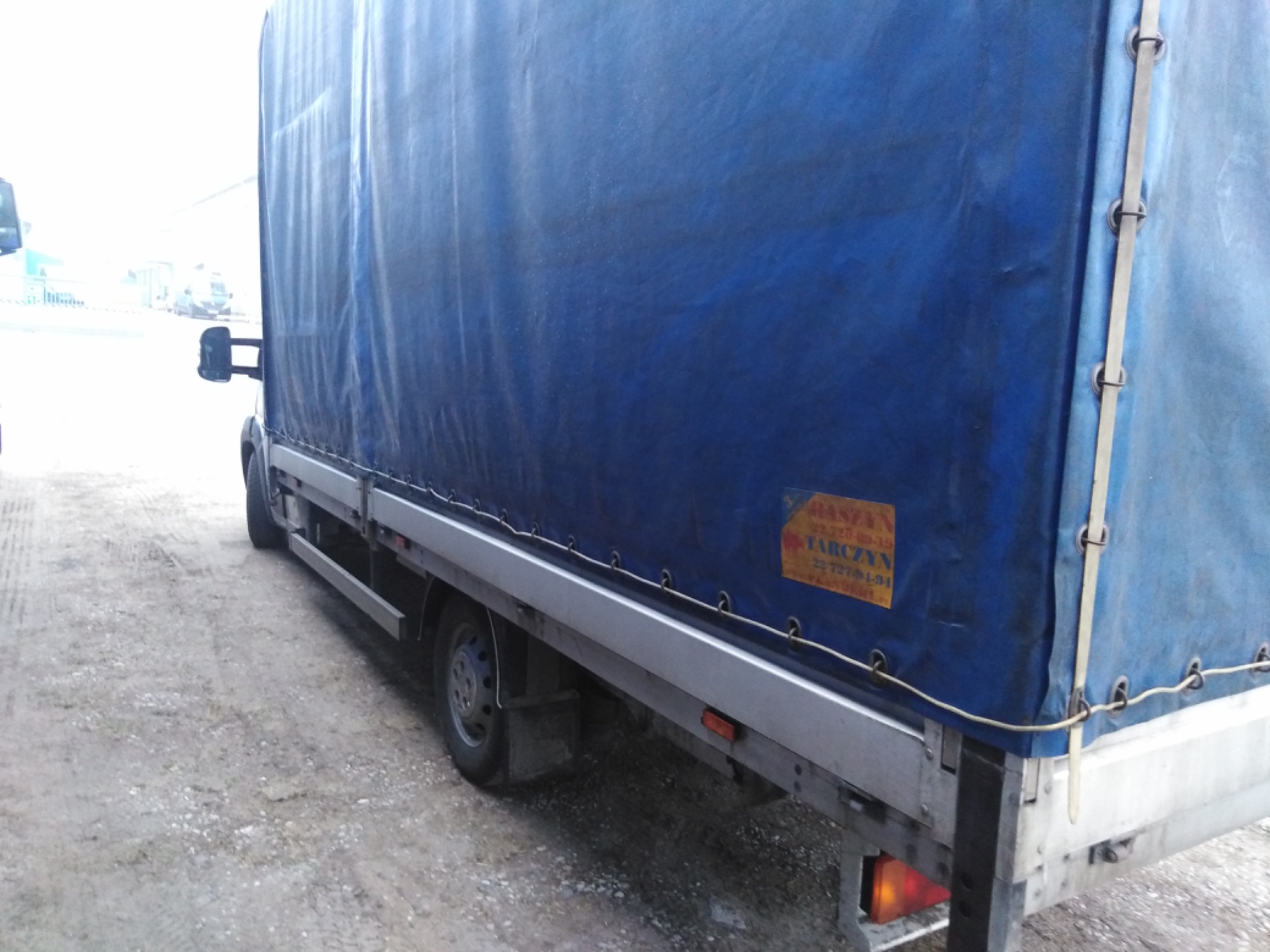 Ciężarówka z niebieską plandeką i żółtą naklejką informacyjną na boku, zaparkowana na szutrowym podłożu.