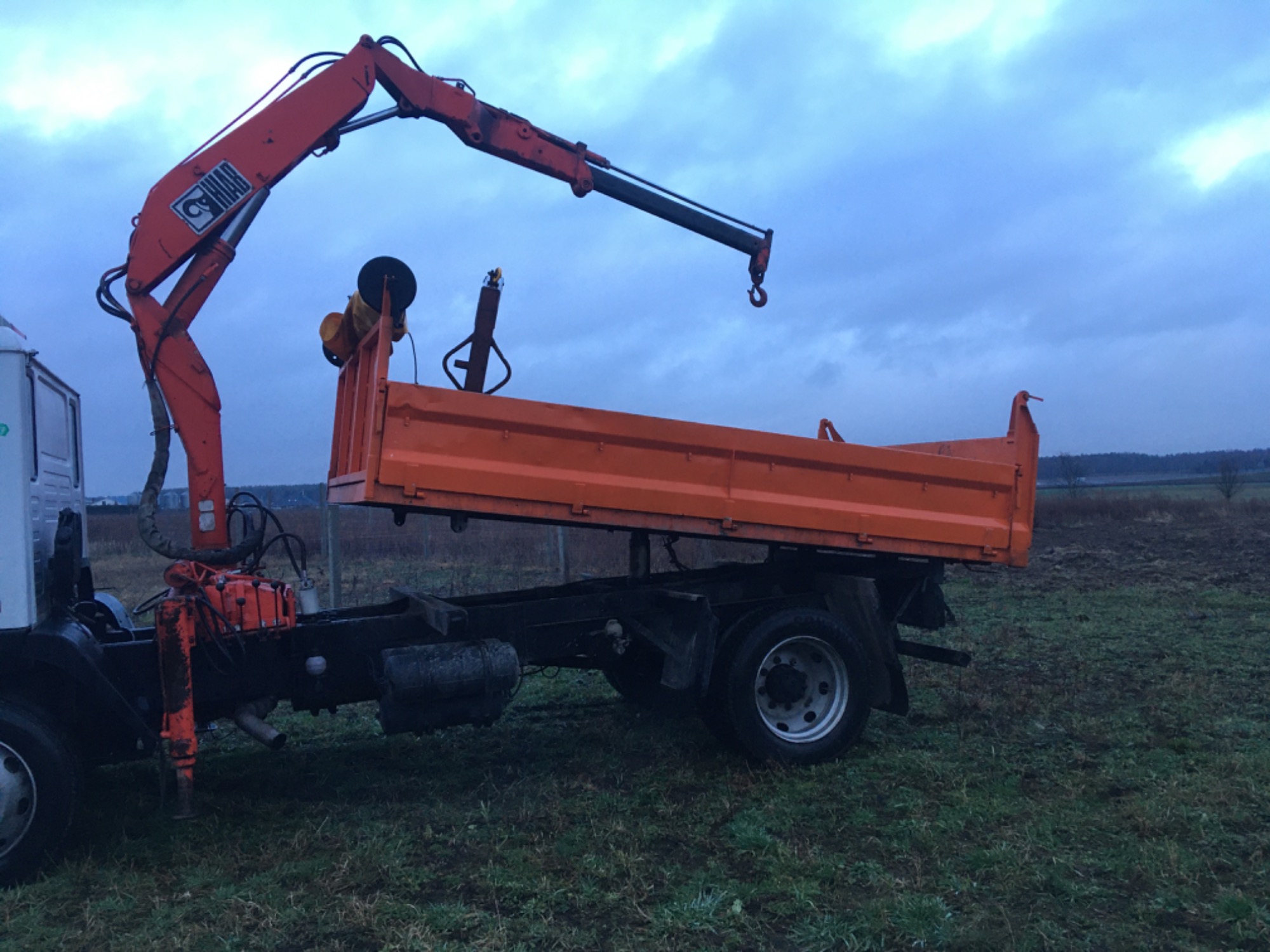 Pomarańczowa wywrotka z dźwigiem HIAB, zaparkowana na trawiastym terenie, pochmurne niebo w tle.