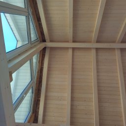 K39 Zakład Konstrukcji Drewnianych - Wnętrze w trakcie budowy: surowe drewno, widoczne elementy konstrukcyjne ścian i stropu, okno dachowe z widokiem na błękitne niebo. Widoczne ocieplenie wokół okna.